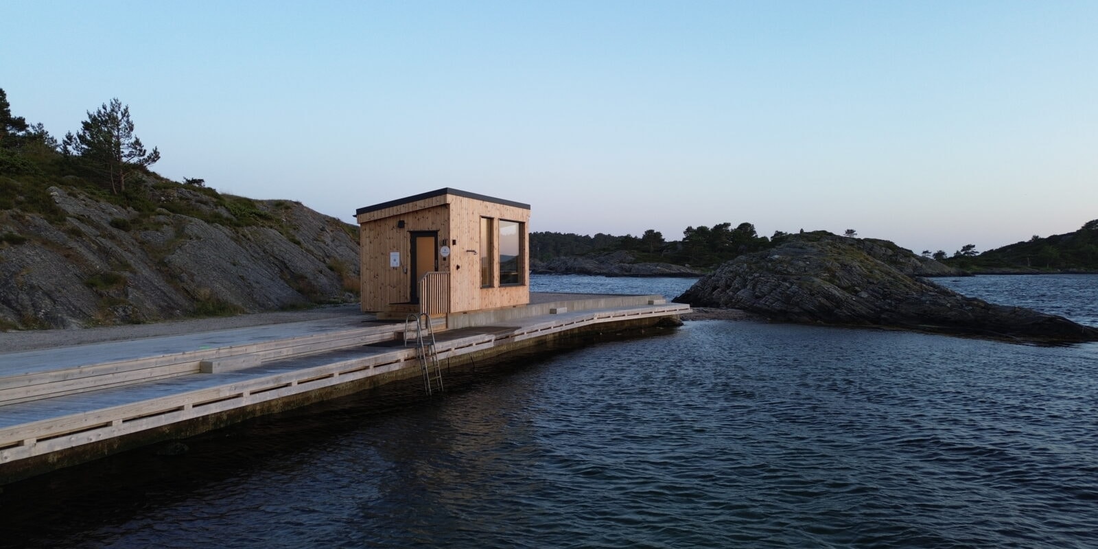 Badstuehyttas moderne, kileformede arkitektur sett fra brygga i klart dagslys – Smeigedag Sauna hos Tilbring i Agder. Rabattkode Tilbring gir 10 % rabatt.