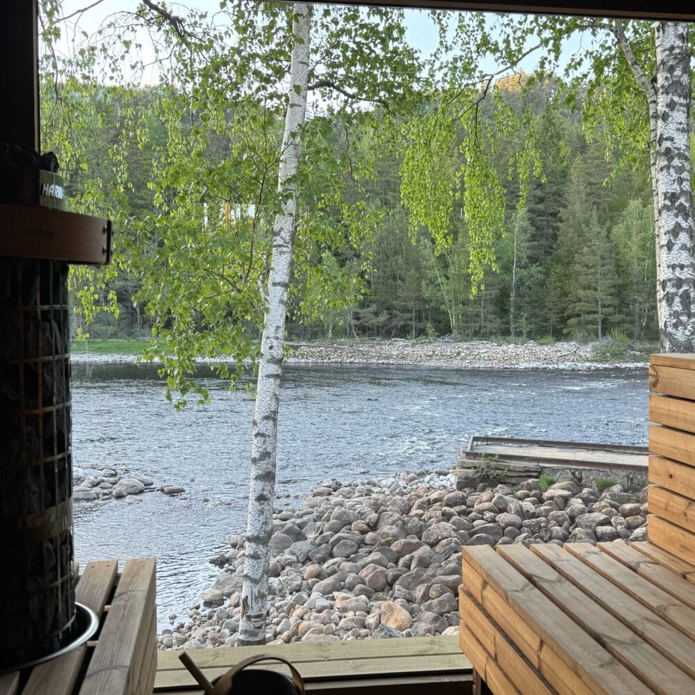 Badstuens interiør med badstueovn og benker, storslått vindusutsikt over vannet mot skogen – Smeigedag Sauna hos Tilbring i Agder. Rabattkode Tilbring gir 10 % rabatt.