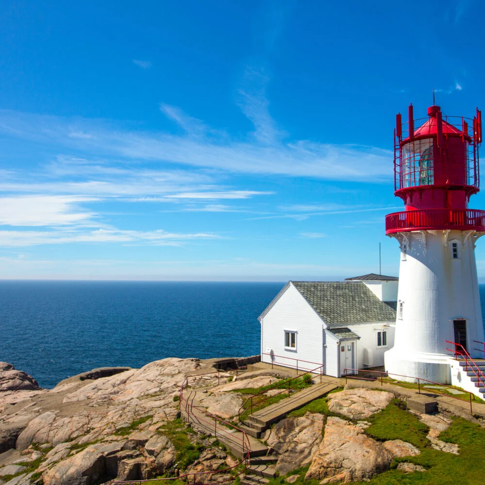 Rødt og hvitt fyrtårn i sol mot blå himmel – Lindesnes Fyr hos Tilbring i Agder.